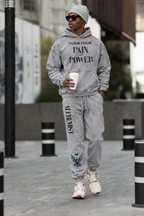A man wearing a matching gray hoodie and jogger sweatpants set with custom text and graphic prints, walking outdoors on a city sidewalk.