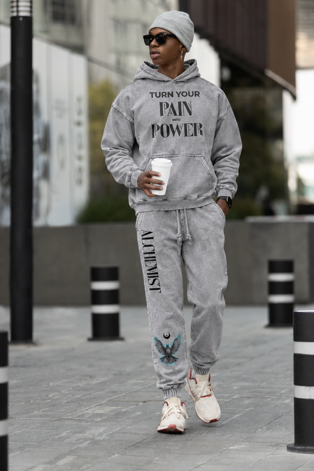 A man wearing a matching gray hoodie and jogger sweatpants set with custom text and graphic prints, walking outdoors on a city sidewalk.