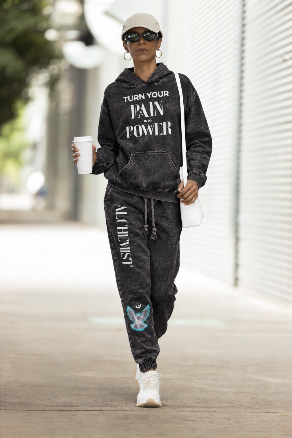 A person walking outdoors wearing a matching black acid-wash hoodie and jogger sweatpants set with white text graphics, white sneakers, a white cap, and sunglasses, while holding a white coffee cup.