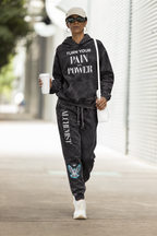A person walking outdoors wearing a matching black acid-wash hoodie and jogger sweatpants set with white text graphics, white sneakers, a white cap, and sunglasses, while holding a white coffee cup.