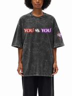 A model wearing an oversized, acid-washed black t-shirt with 'YOU vs. YOU' printed across the chest in red, white, and purple neon-style lettering, paired with black pants against a white background.